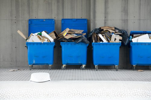 Six blue wheeled rubbish bins are positioned in a row on a concrete pavement against a grey concrete wall. The bins are filled with a variety of waste materials, including cardboard boxes, paper, and plastic packaging, with some overflowing outward. The first bin on the left has its lid open, revealing the contents inside, while the second and third bins are partially covered with black plastic bags containing additional waste. The remaining bins are open or partially covered, displaying a mix of recyclables and general rubbish. A white plastic bag lies on the ground in front of the bins, on the patterned pavement. This scene suggests a commercial waste collection point, possibly in Sanderstead, highlighting the importance of proper rubbish disposal managed by services like Commercial Waste Sanderstead in the local area.