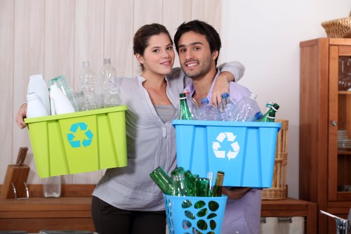 A young man and woman standing indoors in a room with light-colored wooden paneling and shelving, each holding a plastic recycling bin filled with empty glass bottles. The woman holds a bright green bin with a recycling symbol on the front, containing mostly clear and light green glass bottles. The man holds a blue bin with a recycling symbol, also filled with clear glass bottles, some of which are leaning against the sides. In front of them, a third smaller blue container is filled with dark green glass bottles, positioned on a wooden surface or countertop. The background includes a wooden shelving unit with baskets and decorative items, and the lighting is soft and natural, emphasizing the clean, organized scene related to waste separation and recycling practices consistent with rubbish collection services in the Sanderstead area.