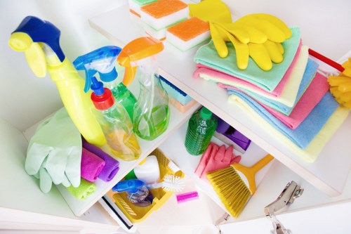 A photograph showing a collection of cleaning supplies on a white kitchen or utility room surface, including spray bottles with colored liquids, rubber gloves in various colors, cleaning cloths and sponges stacked and folded, a yellow-handled scrub brush, and a dustpan with a small brush. The background features a blurred view of additional cleaning cloths with bright colors such as pink, green, and blue, placed on a shelf or countertop. The items are arranged somewhat disorderly, indicating they are ready for use or have been recently used, and they are all part of household or commercial cleaning in a domestic or light commercial setting. Commercial Waste Sanderstead offers rubbish collection and waste removal services which include maintaining cleanliness and disposal of cleaning-related waste, suitable for properties in the Sanderstead area, close to the postcode region inferred from the page context. This detailed visual layout reflects typical cleaning equipment used in premises where rubbish removal by professional services would be applicable.