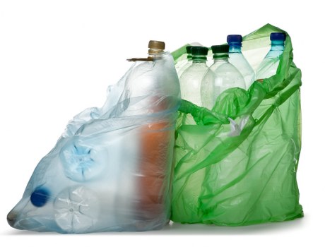 A group of used plastic bottles and containers, including a clear bottle with a beige cap, a transparent bottle with a blue cap, and a green basket liner, arranged on a plain white background. The bottles vary in shape and size, with some appearing crumpled or squeezed, indicating they have been used. The green bag or liner holds additional bottles and possibly other lightweight plastic items, and is partially open, allowing visibility of the bottles inside. The materials have a smooth, glossy texture typical of recycled plastic, with some bottles showing slight creases or dents from prior use. The scene focuses on these typical items collected for rubbish removal services, reflecting a domestic or commercial waste collection scenario. The image has no other objects or environmental context visible, but it relates to waste disposal and recycling efforts often handled by services such as those offered by Commercial Waste Sanderstead in the Sanderstead area.