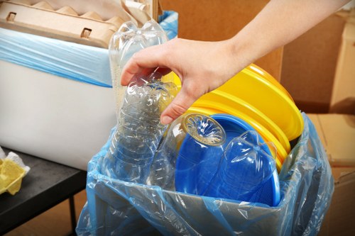 A close-up of a person's hand placing a clear plastic water bottle into a blue and transparent rubbish bag lined with a blue plastic liner. The background shows a black surface, possibly a table, with a cardboard box in the upper left corner and several brightly coloured plastic bowls stacked inside the bag, including yellow and blue ones. The scene appears to be set indoors, likely in a utility or storage area where waste is being collected or prepared for removal. The focus is on the process of disposing of recyclable plastic containers, relevant to commercial waste removal services in Sanderstead. The image emphasizes the sorting of waste materials for effective rubbish collection and disposal, consistent with professional waste management practices provided by Commercial Waste Sanderstead.
