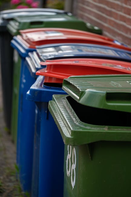 A row of large outdoor wheelie bins lined up along a paved pathway next to a brick wall, with the first in the row being a green bin featuring a textured lid and partial white recycling symbols on its