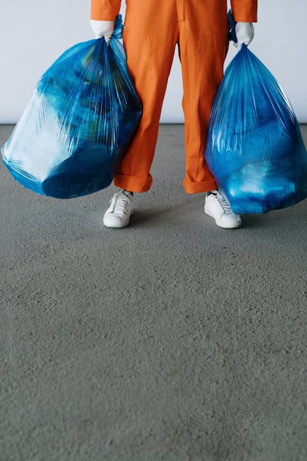 A person standing on a grey pavement, holding two large blue plastic rubbish bags filled with waste, one in each hand. The individual is wearing orange work overalls, white sneakers, and is positioned