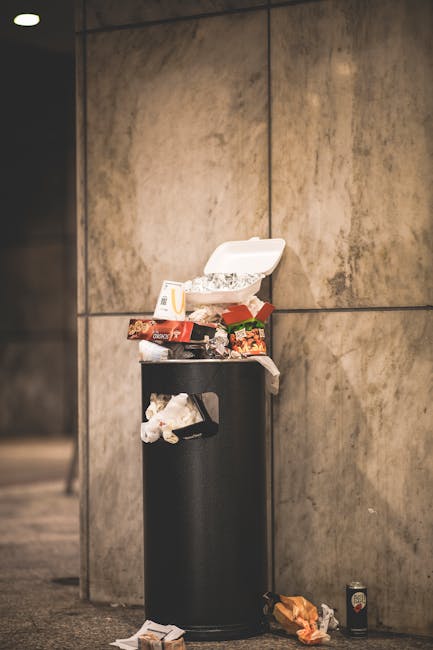 A small blue pickup truck parked on the side of the road, loaded with a large collection of bulky waste items, including cardboard boxes, black plastic bags, and miscellaneous discarded materials. The