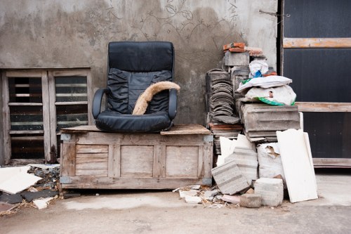 In an outdoor, urban setting, there is a black leather office chair placed atop a wooden crate, with its armrests and cushioned back visible, positioned against a weathered, beige wall that shows signs of wear and grime. To the right of the chair, a pile of construction debris and waste materials is stacked on the ground, including irregularly shaped concrete blocks with textured surfaces and visible aggregate, some with white or grey finishes, along with a few bricks and fragments of plaster. A small cardboard box or package is also part of the debris. The surrounding environment includes a concrete pavement and a metal or wooden structure visible in the background, hinting at an industrial or back alley area. The scene captures a typical example of waste accumulation that a rubbish removal service, such as Commercial Waste Sanderstead, may handle as part of their local junk disposal and clearing work in the Sanderstead area.