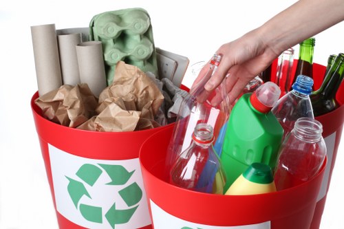 A close-up view of a red waste collection bin with a recycling symbol, filled with various waste materials including several cardboard toilet paper tubes, crumpled brown paper, and green plastic egg cartons. Adjacent to this, another similar red bin contains plastic bottles and containers in different sizes and colors such as transparent, green, and red, with some having caps still attached. The bins are situated on a plain, white background, emphasizing the variety of household waste types collected for disposal or recycling, typical of rubbish removal services provided by companies like Commercial Waste Sanderstead. The image highlights the importance of separating recyclable plastics from other waste and demonstrates the diversity of materials involved in domestic rubbish management, reflective of local efforts to maintain cleanliness and environmental standards within the Sanderstead area, postcode range inferred from the webpage context.