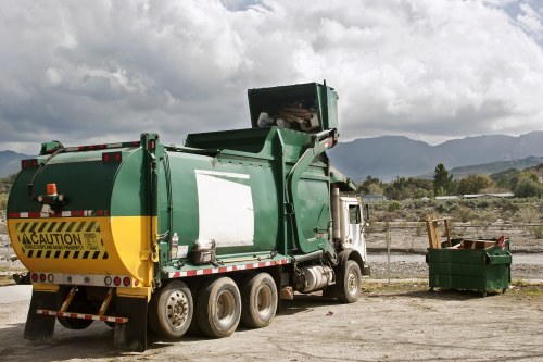 A large green waste collection truck parked outdoors on a gravel surface, with a black and yellow caution stripe on the rear. The truck features a large, open-top compactor unit at the back, designed for gathering and compressing rubbish. Nearby, a small dark green skip bin filled with mixed waste materials is positioned on the right side of the truck. The scene is set against a backdrop of distant hills or mountains under a partly cloudy sky, indicating a rural or semi-rural area possibly near Sanderstead. The environment appears to be a waste disposal or collection site, with no other vehicles or structures in immediate view, emphasizing the operational aspect of rubbish removal services. The lighting suggests daylight hours, highlighting the textures of the gravel, the truck’s metallic surface, and the waste bin's contents, which include loose debris and recyclable materials.